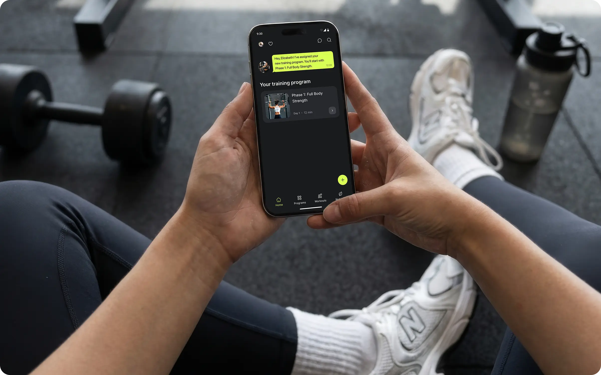 Persona en un gimnasio consultando el programa de entrenamiento asignado y un mensaje del entrenador en la app Ymove en iPhone